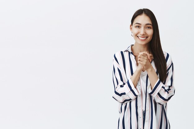 Girl looking with caring and admirable eyes, being thrilled and charmed with pleasant gift, clasping hands over chest and smiling with happiness, standing in striped blouse over gray wall