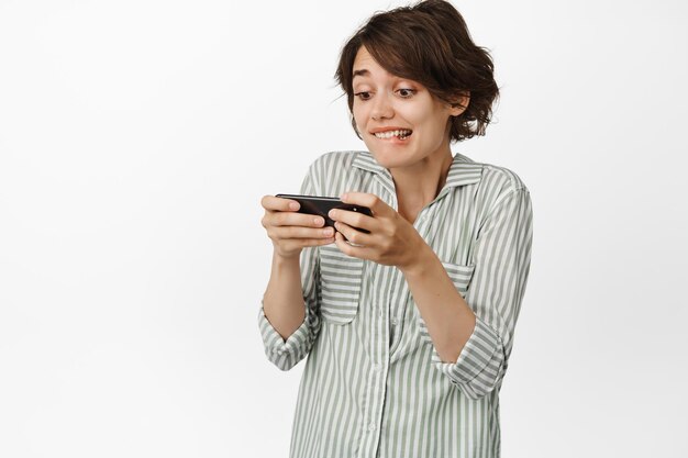 Girl looks excited at smartphone screen, watching on mobile phone, playing video games, standing against white background