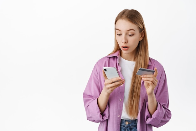 Girl looks surprised at smartphone screen while paying for online shopping order, holding credit card, reading app notification on mobile phone during purchase, standing over white wall