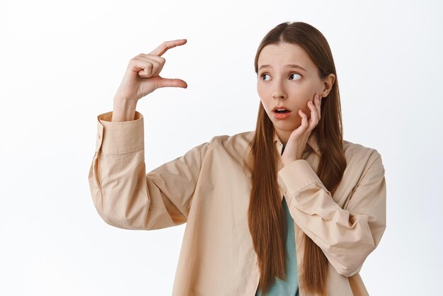 Girl looks troubled at small tiny size showing little thing and staring worried at product on copy space standing against white background