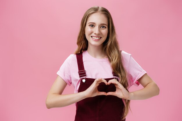 Girl loves family. Kind charming young woman in overalls with small tattoo on arm showing heart gesture over body and smiling lovely at camera expressing tender and cute attitude over pink wall.