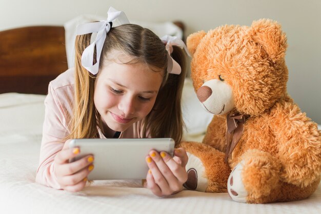 Girl lying on bed with teddy bear looking at mobile phone