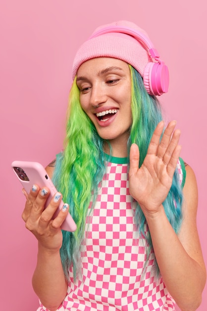  girl makes greeting gesture waves hand to camera and says hello uses wireless headphones and modern smartphone enjoys video conversation with friend poses on pink 