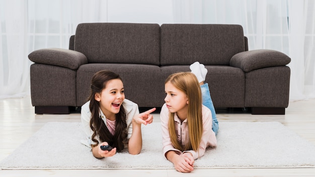 Девушка шутит над подругой, лежащей на ковре у себя дома