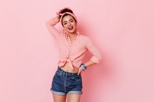 Девушка в массивных серьгах, розовой рубашке и джинсовых шортах позирует на розовом пространстве с поднятой рукой.
