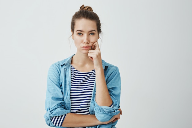 Girl need to find answer. Charming young caucasian woman with bun hairstyle holding fingers on cheek and chin, looking seriously and troubled , thinking or deciding something in mind