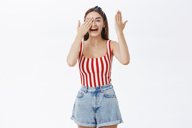 Girl opens eyes seeing amazing surprise laughing and smiling broadly from joy and amazement holding palm on eye peeking, amused and enthusiastic over gray wall