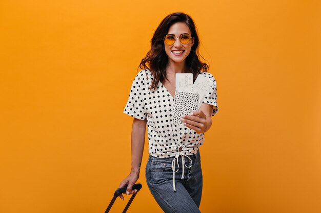 Девушка в оранжевых очках держит билеты и чемодан. Темноволосая взрослая женщина в клетчатой рубашке позирует и улыбается на изолированном фоне.