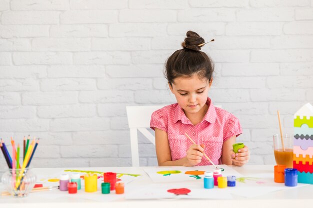 A girl painting with watercolors on table against white brick wall