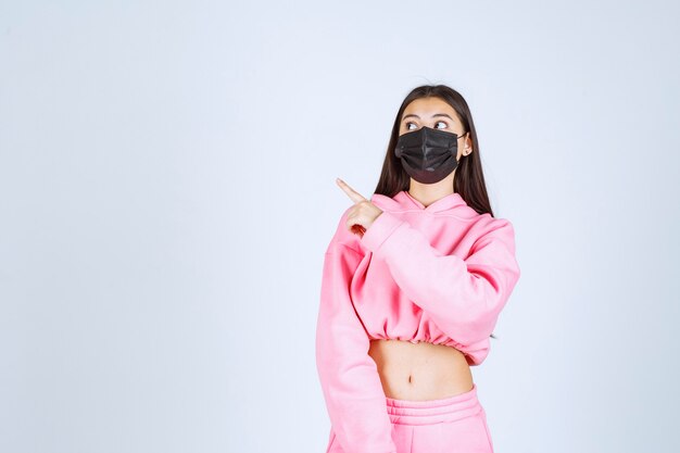 Girl in pink pajamas and black mask pointing left side. 