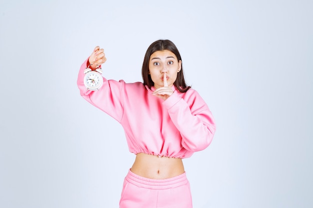 Girl in pink pajamas holding an alarm clock and asking for silence. 