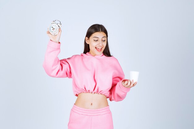 Girl in pink pajamas holding an alarm clock and a cup of coffee. 
