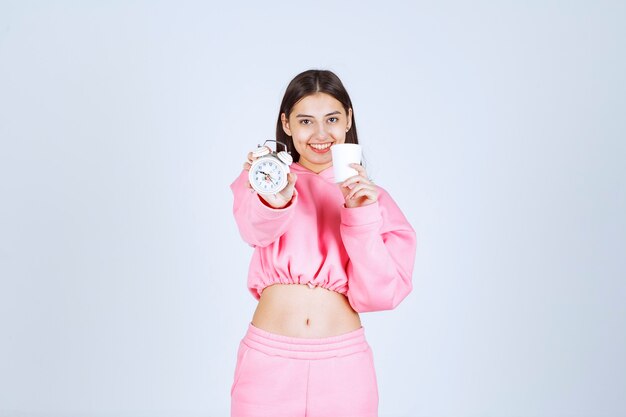 Girl in pink pajamas holding an alarm clock and a cup of coffee. 