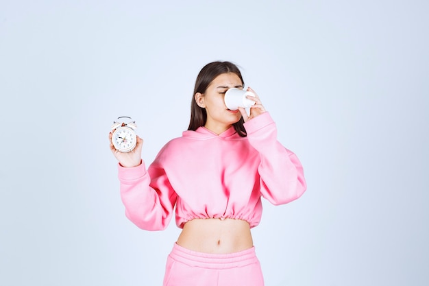 Girl in pink pajamas holding an alarm clock and drinking a cup of coffee. 