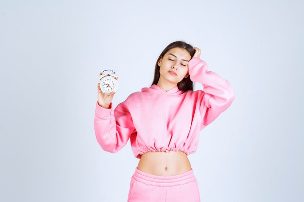 Girl in pink pajamas holding an alarm clock and looks sleepy. 