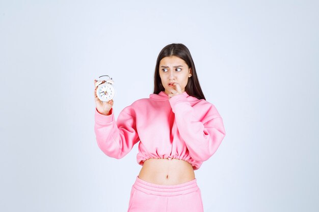 Girl in pink pajamas holding an alarm clock and looks thoughtful and confused. 