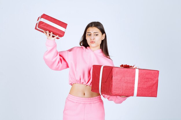 Девушка в розовой пижаме держит большие и маленькие красные подарочные коробки и выглядит недовольной.