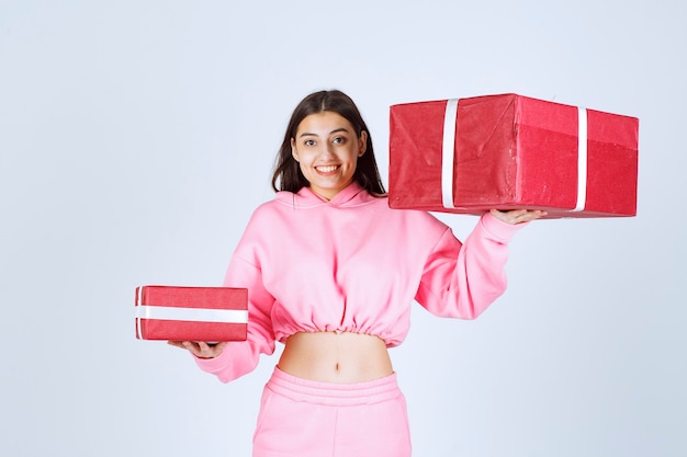 Girl in pink pajamas holding big and small red gift boxes and smiling. 