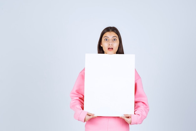 Girl in pink pajamas holding a blank square presentation board in front of her. 