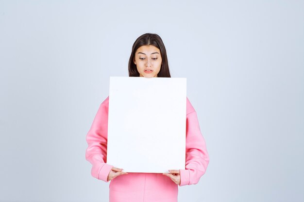 彼女の前に空白の正方形のプレゼンテーションボードを保持しているピンクのパジャマの女の子。