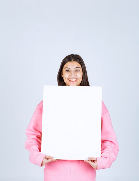 Girl in pink pajamas holding a blank square presentation board in front of her. 