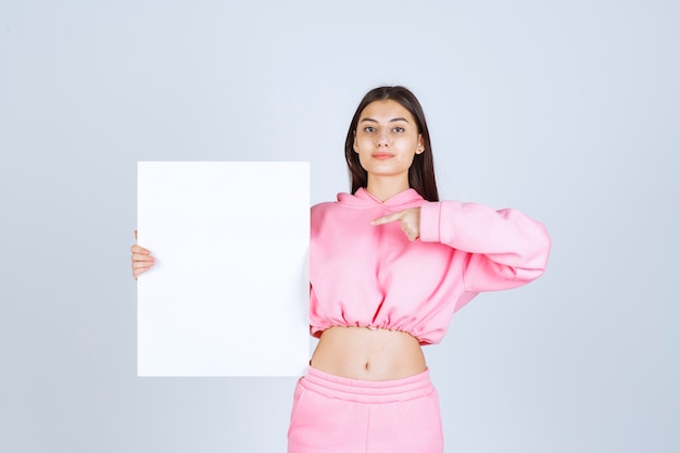 Girl in pink pajamas holding a blank square presentation board and pointing at it. 