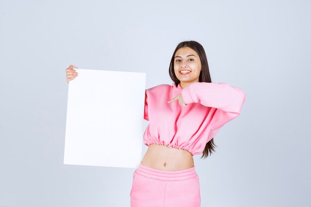 Girl in pink pajamas holding a blank square presentation board and pointing at it. 