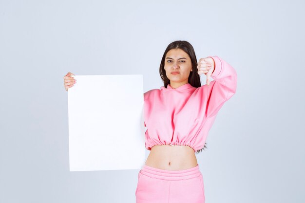 Girl in pink pajamas holding a blank square presentation board and showing thumb down. 