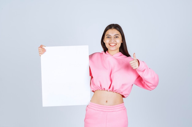 Girl in pink pajamas holding a blank square presentation board and showing thumb up sign.