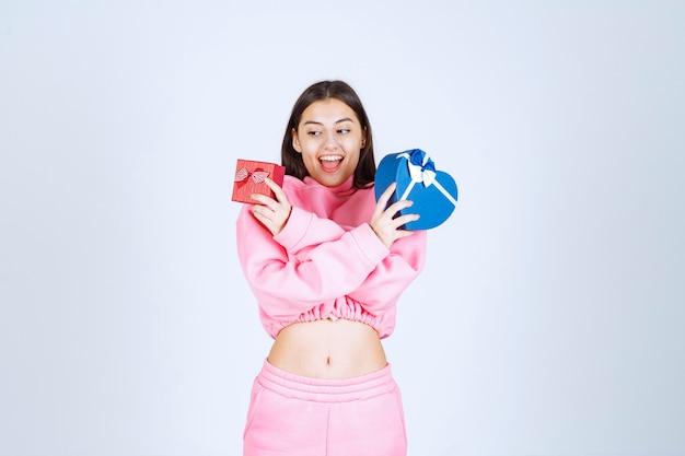 Girl in pink pajamas holding red and blue heart shape gift boxes in both hands. 