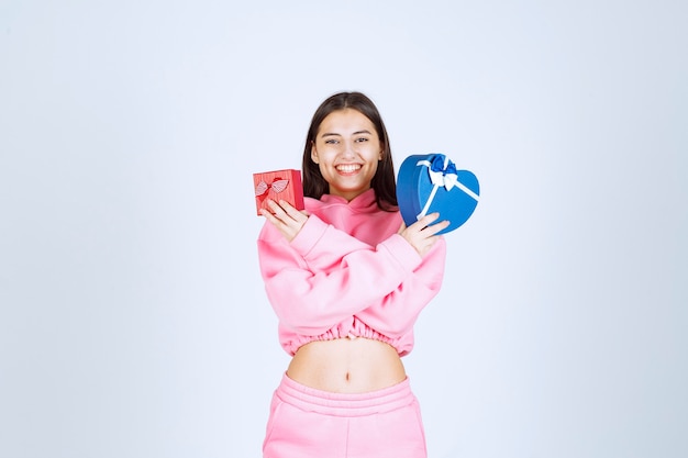 Girl in pink pajamas holding red and blue heart shape gift boxes in both hands. 