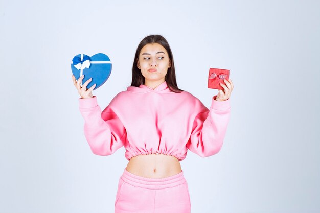 Girl in pink pajamas holding red and blue heart shape gift boxes and making choise between them. 