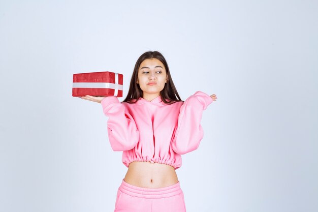 Girl in pink pajamas holding a red rectangular gift box and looks indifferent. 