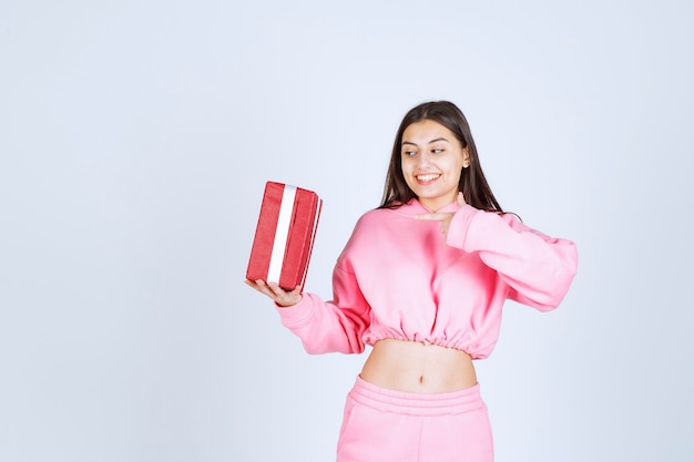 Girl in pink pajamas holding a red rectangular gift box and looks satisfied. 