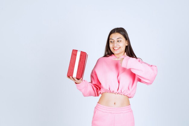 Девушка в розовой пижаме держит красную прямоугольную подарочную коробку и выглядит довольной.