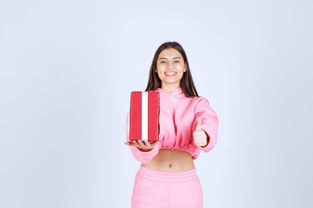 Girl in pink pajamas holding a red rectangular gift box and looks satisfied. 