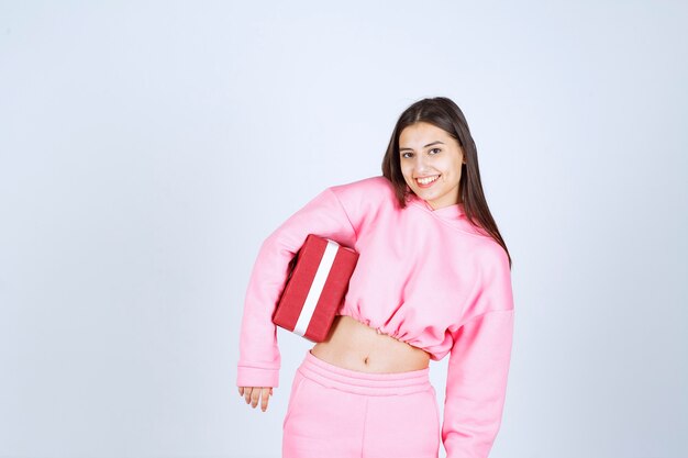 Girl in pink pajamas holding a red rectangular gift box and looks satisfied. 