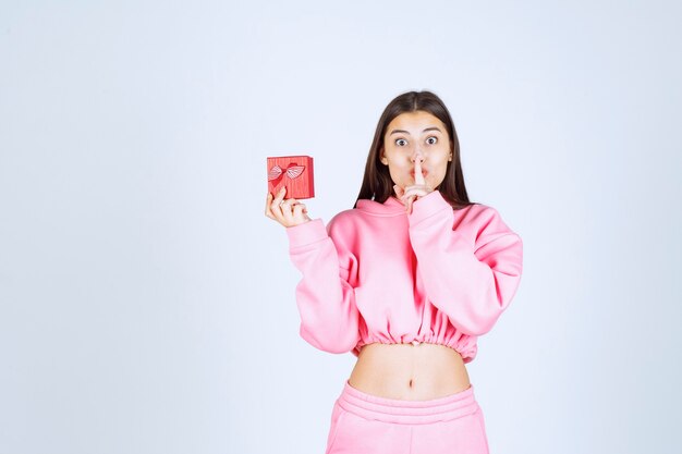 Girl in pink pajamas holding a small red gift box and asking for silence. 