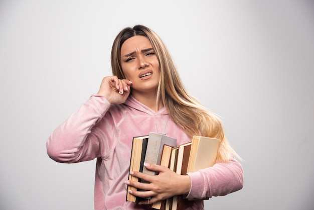 Girl in pink swaetshirt holding a stock of books and feels tired.