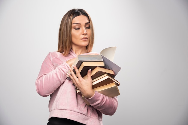 Girl in pink swaetshirt holding a stock of books, opening one on the top and reading it