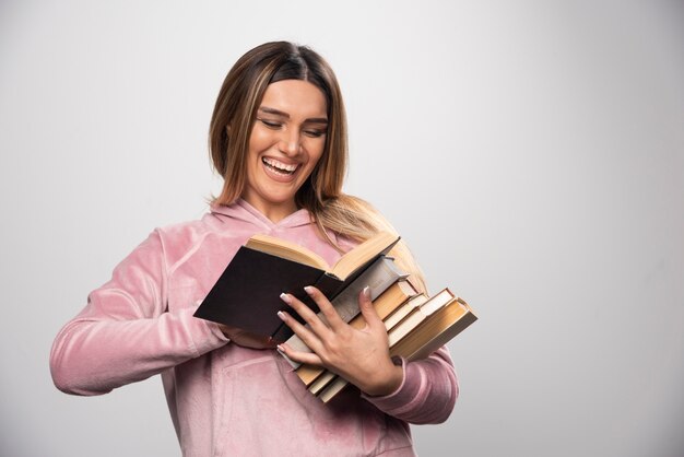 Girl in pink swaetshirt holding a stock of books, opening one on the top and reading it