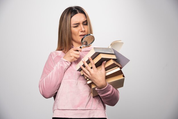 Девушка в розовой рубашке держит стопку книг и пытается прочитать верхнюю с лупой