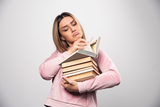 Girl in pink sweatshirt holding a stock of books, opening one on the top and reading it.