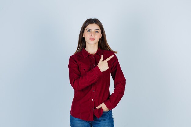 girl pointing at upper right corner, holding hand in pocket in burgundy shirt and looking confident.