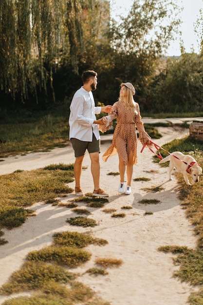 Girl in polka-dot dress and her boyfriend in khaki shorts are walking large white dog in park.