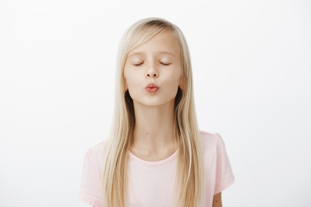 Girl practising near mirror how to kiss. Portrait of cute fashionable young girl with blond hair, folding lips and closing eyes while waiting for a kiss