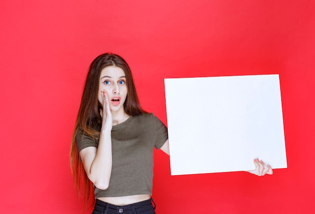 Free photo girl presenting a square info board and looks surprized and confused.