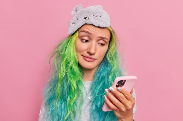  girl reads information on smartphone looks with unhappy expression gets message from formal boyfriend wears sleepmask on forehead isolated on pink 