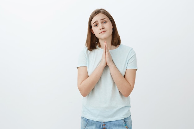 Girl really needs help asking it with cute angel face. Tender and attractive young woman in t-shirt and jeans tilting head smiling gentle, holding hands in pray while begging favor