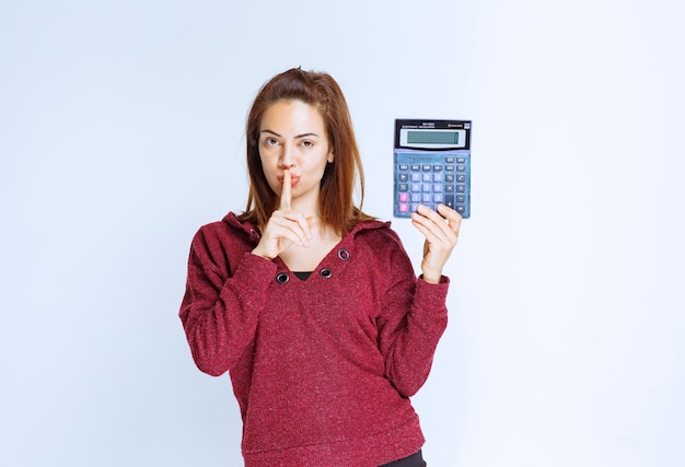 Girl in red jacket calculating something on a blue calculator, showing the result and asking for silence.